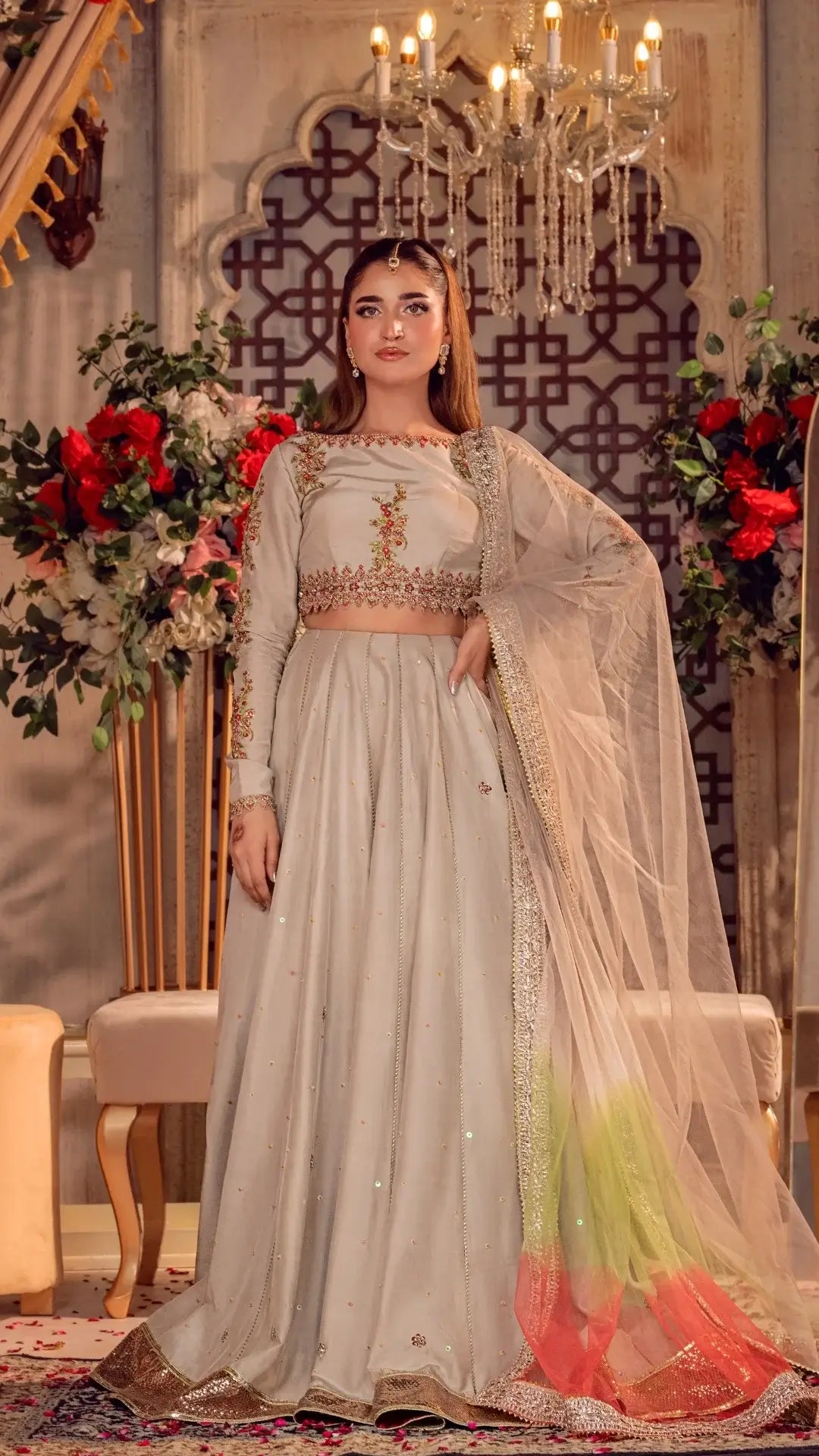 Woman in an elegant ivory Pakistani formal dress with floral embroidery and net dupatta, styled for a wedding or party, standing in a lavish indoor setting with chandeliers and floral decor.