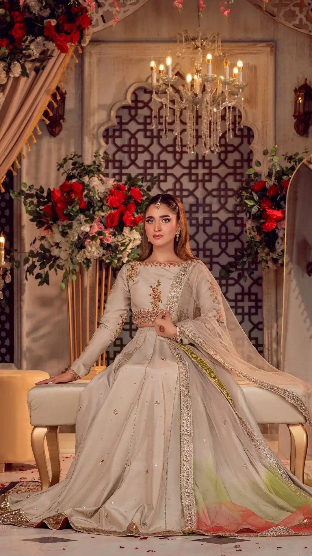 Woman in an embroidered traditional outfit sitting in a decorated room with flowers and a chandelier.
