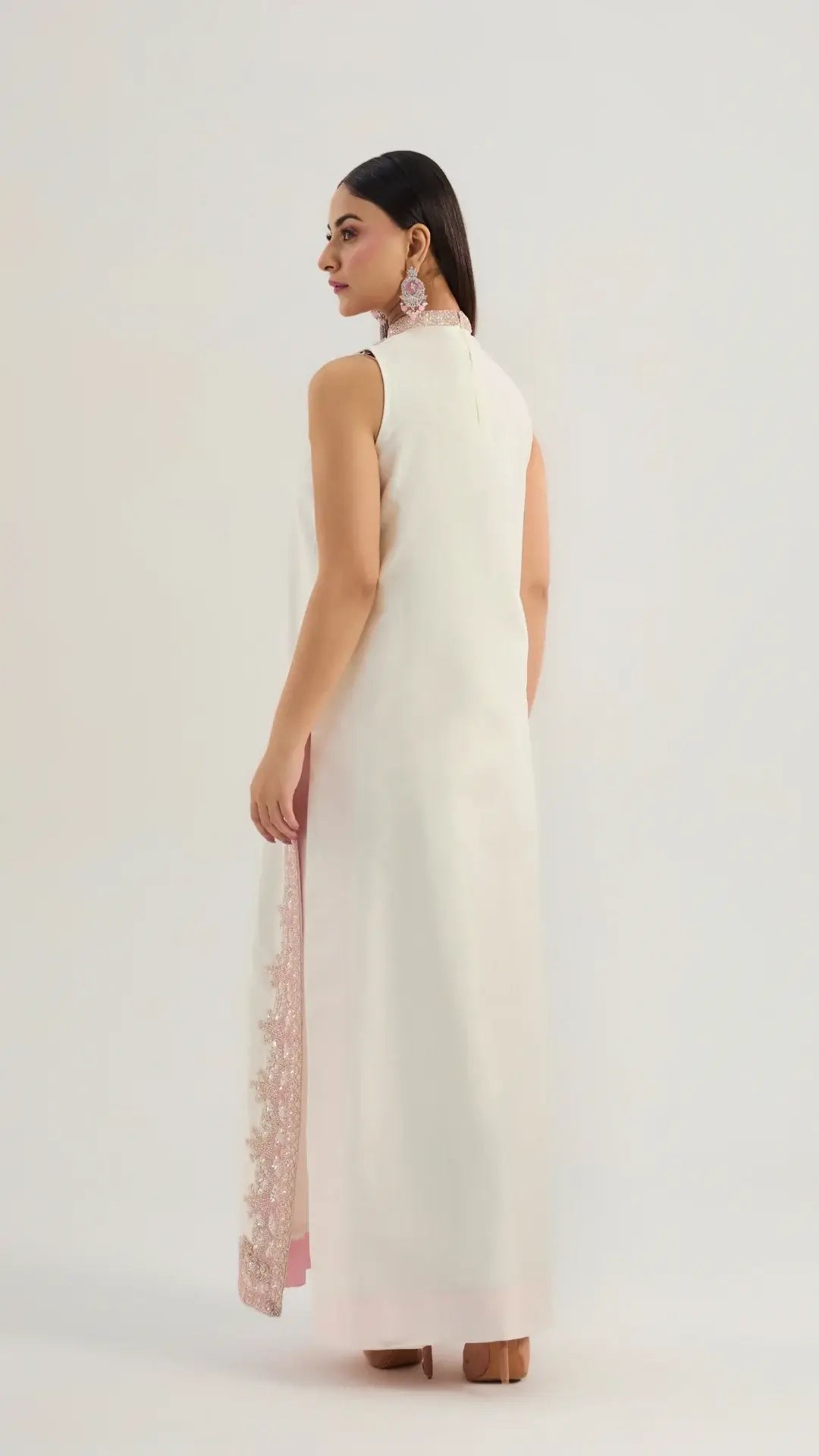 Woman wearing a white sleeveless dress with a high neckline and lace details on a plain background