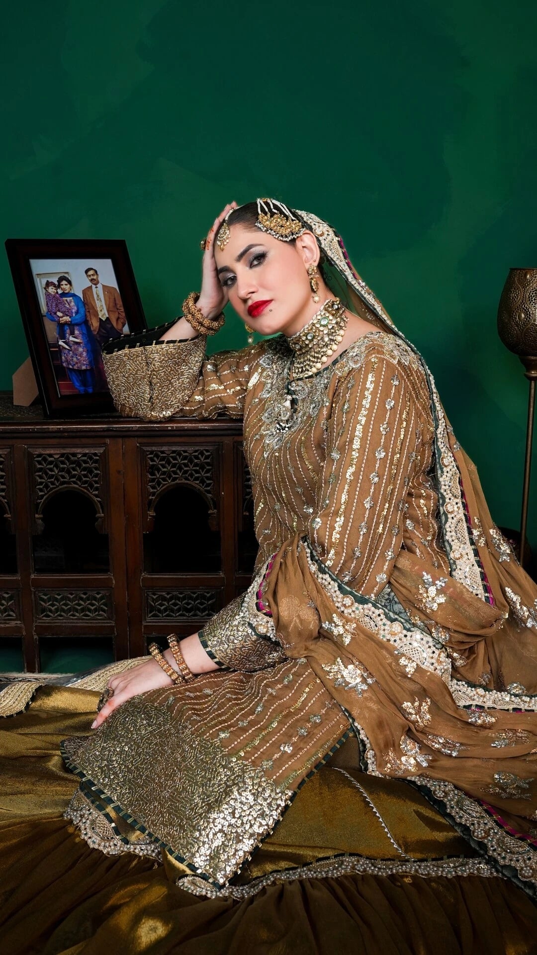 Woman in traditional embroidered outfit with jewelry against a green wall.