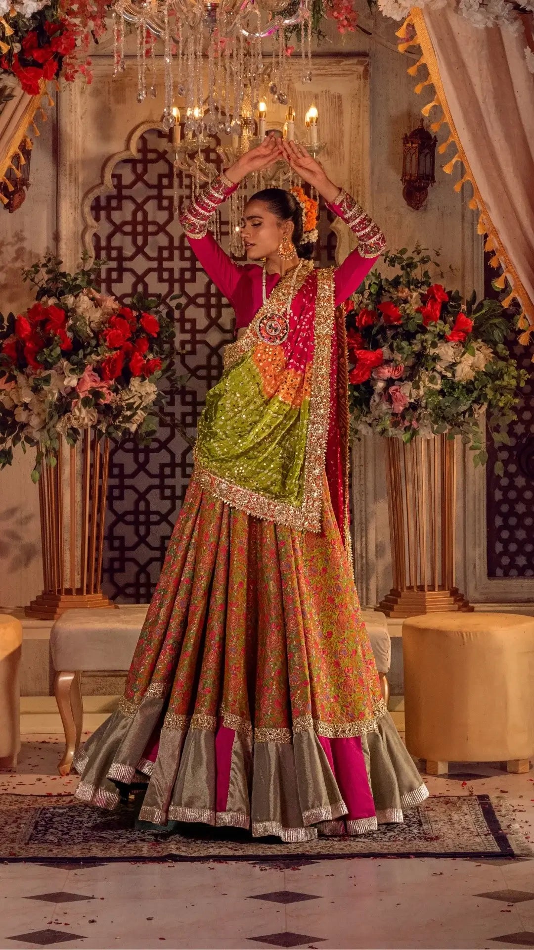 Woman in a vibrant Pakistani formal lehenga with floral patterns and embellished dupatta, standing in an ornate room with floral decor.