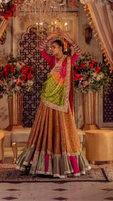 Woman in a vibrant Pakistani formal lehenga with floral patterns and embellished dupatta, standing in an ornate room with floral decor.