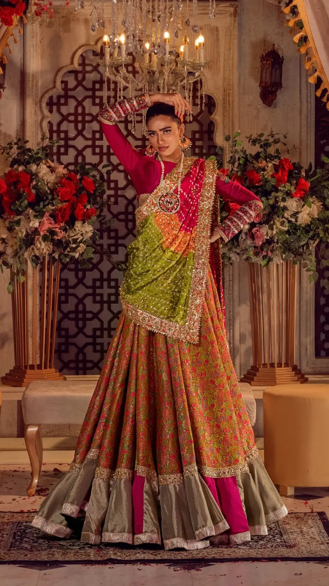 Woman in vibrant formal Pakistani dress with gold embellishments under chandelier, floral backdrop