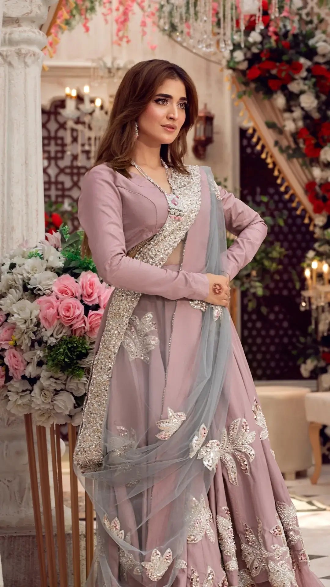 Woman wearing a mauve embroidered Pakistani formal dress with dupatta, floral decor in background
