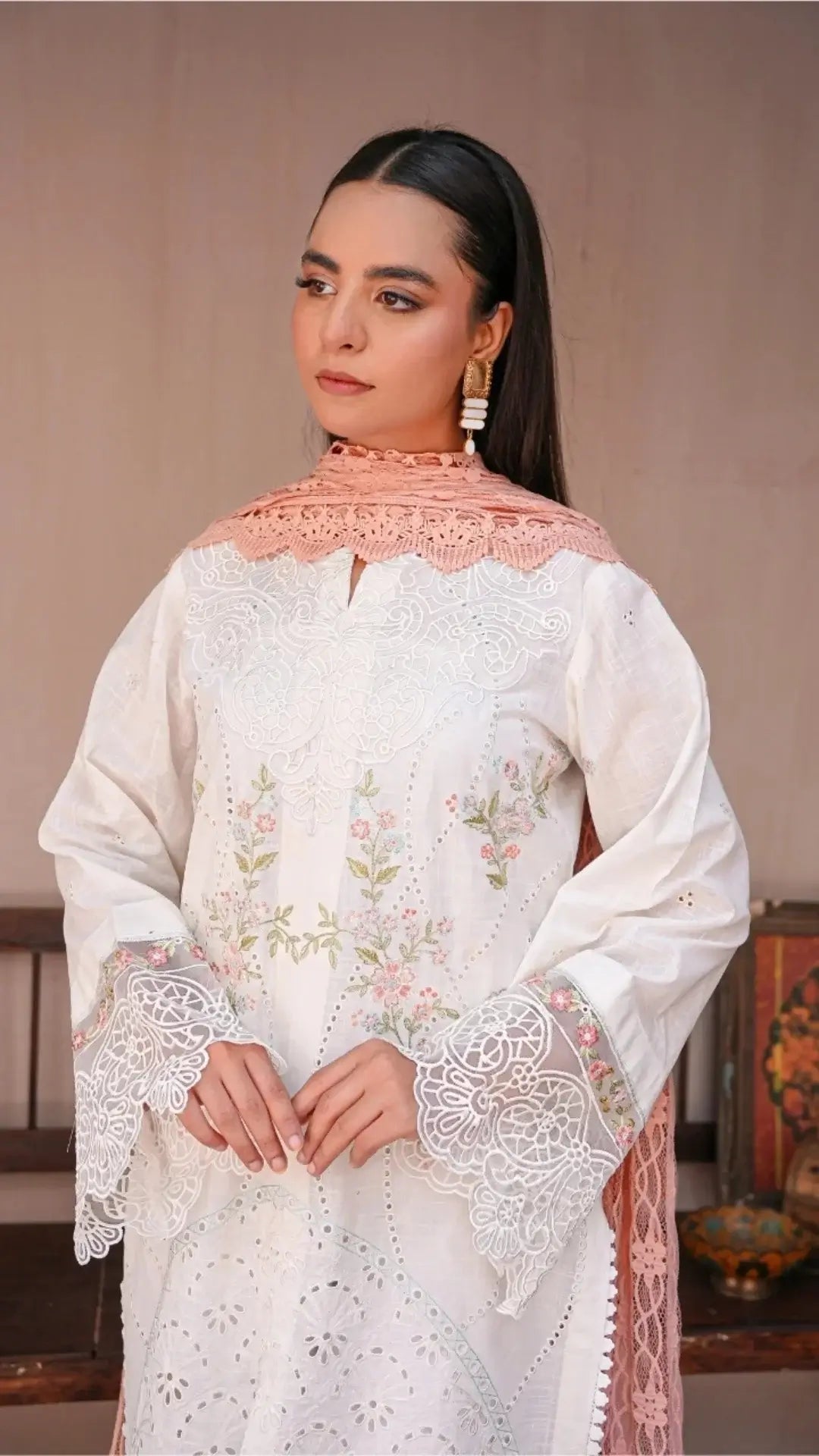 Woman wearing a white embroidered traditional outfit with a pink scarf against a neutral background
