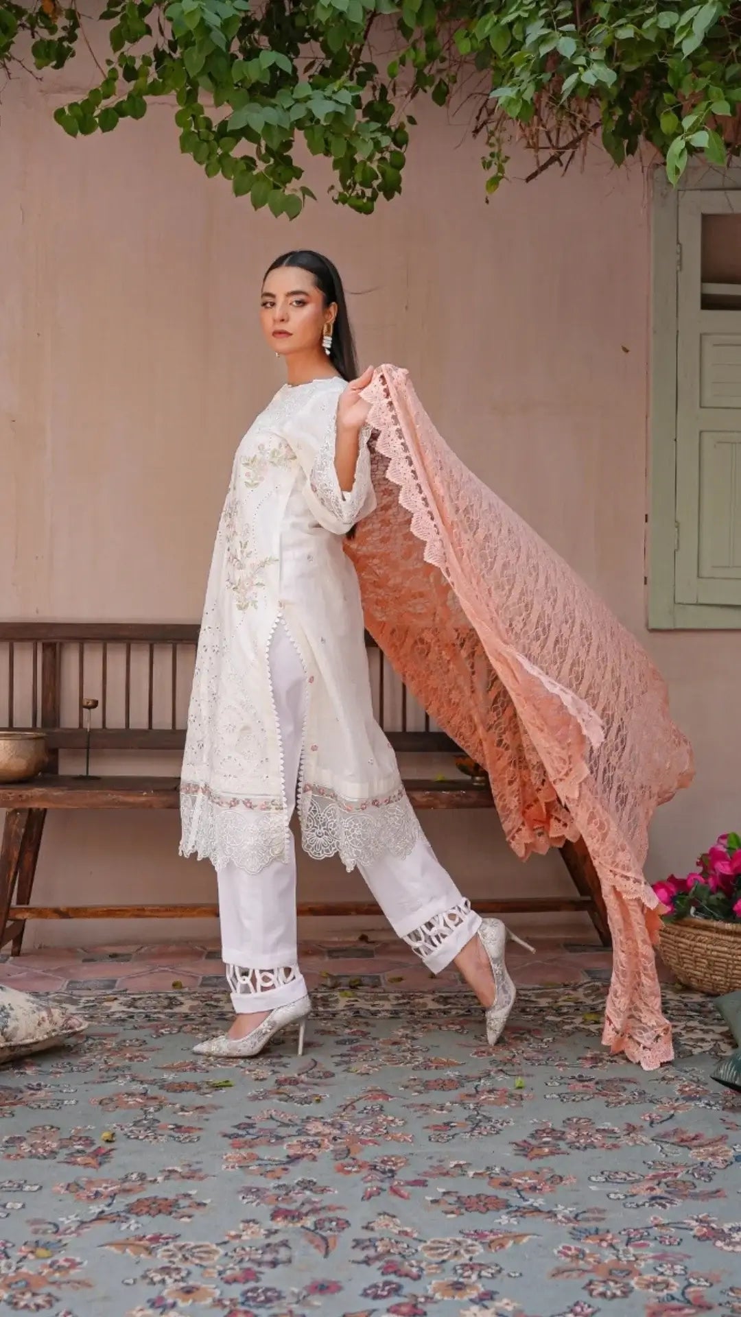 Woman in a white traditional outfit with a pink lace dupatta standing in a rustic setting.