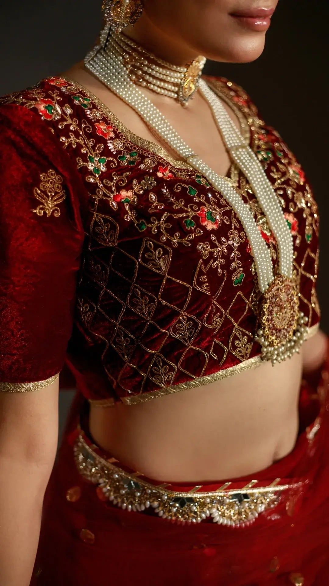 Red traditional outfit with intricate gold embroidery and pearl necklace on a dark background