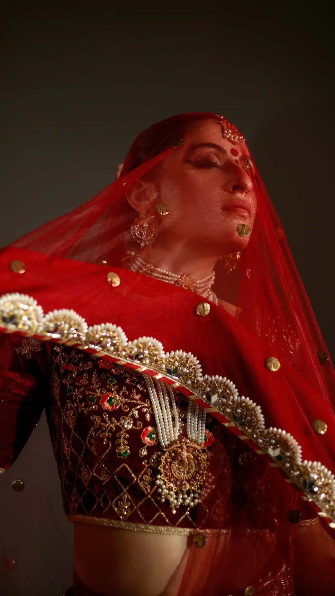 Woman in traditional red and black embroidered outfit with gold jewelry against a dark background