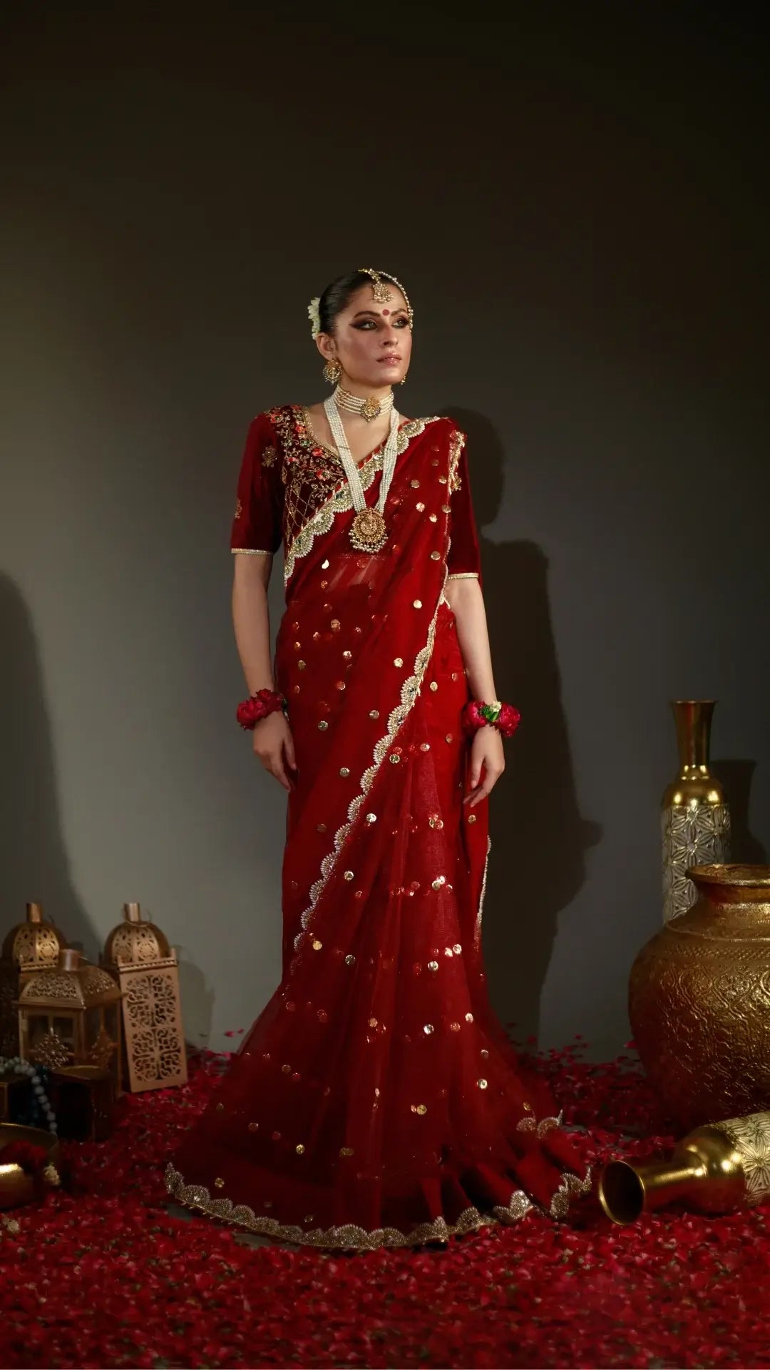 Woman in a red saree with gold details standing against a dark background with decorative elements.