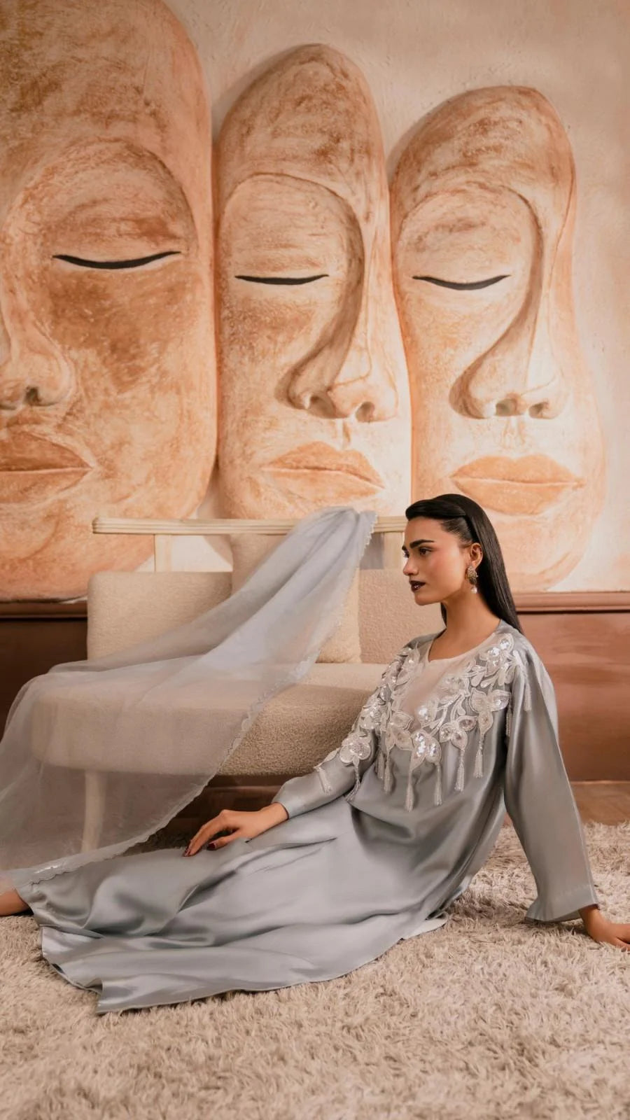 Woman in a long, light gray dress sitting in front of a wall with Buddha statues.