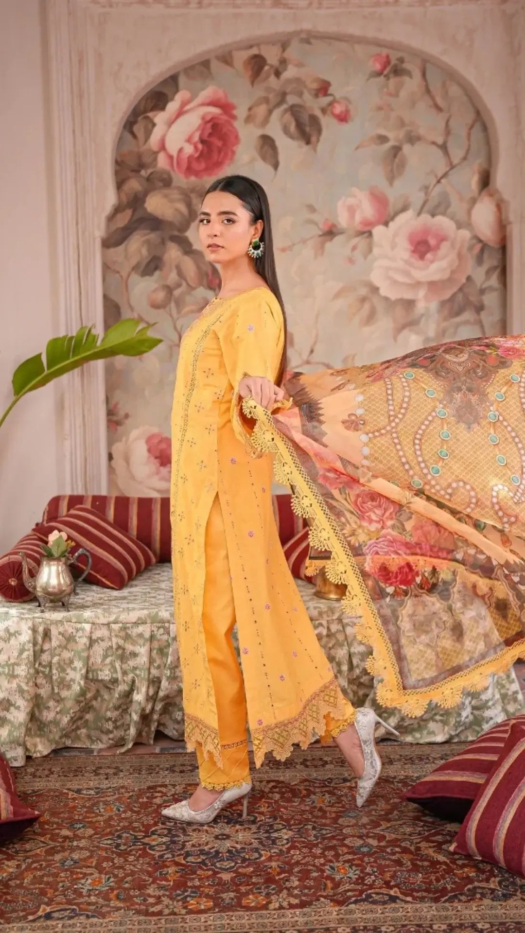 Woman in a yellow traditional outfit standing in a decorated room with floral wallpaper.