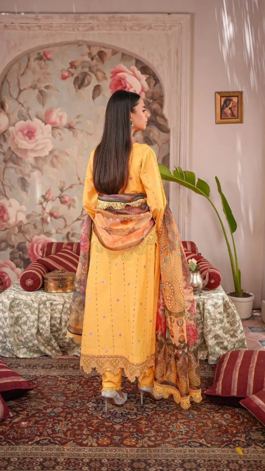 Woman in a yellow traditional outfit standing in a decorated room with floral wallpaper and red furniture.