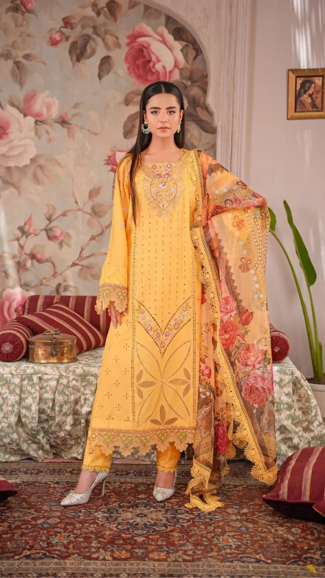Woman in a yellow embroidered outfit standing in a decorated room with floral wallpaper.